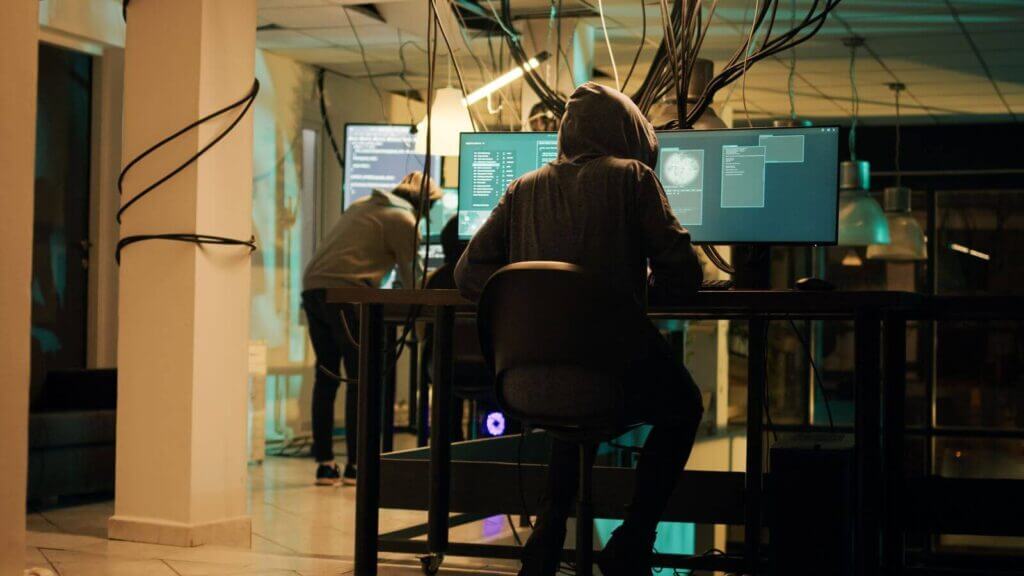 Two individuals in hoodies work at computer stations connected to multiple monitors in a dimly lit room, surrounded by exposed cables and dim lighting.