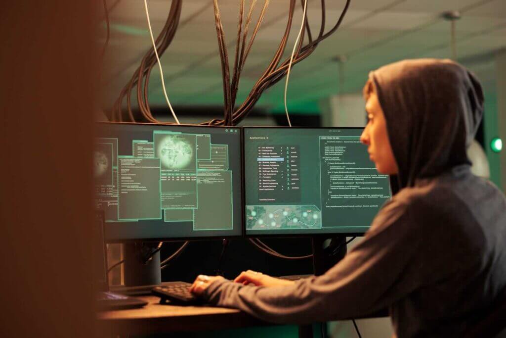 A person in a hoodie is using a computer with multiple monitors displaying complex data and code in a dimly lit room.