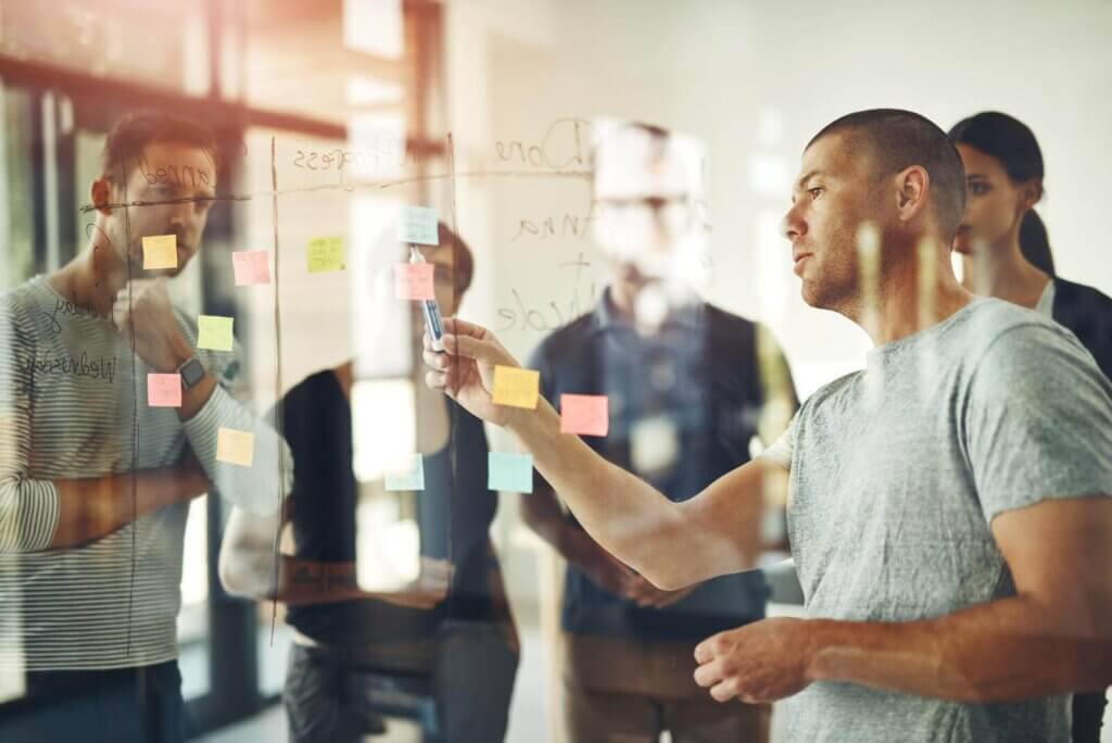 A group of people collaboratively discussing notes on a glass wall covered with sticky notes in an office setting.