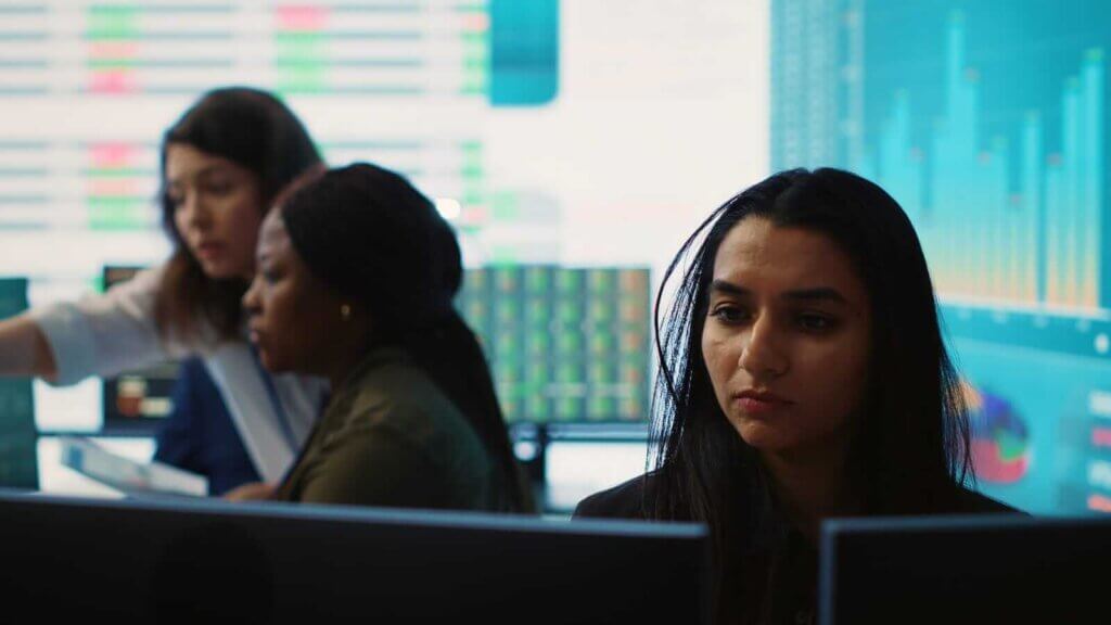 A group of people work in a busy office, focusing on computer screens displaying data and charts.