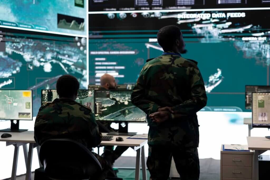 Military personnel in a control room monitor multiple screens displaying data and maps.