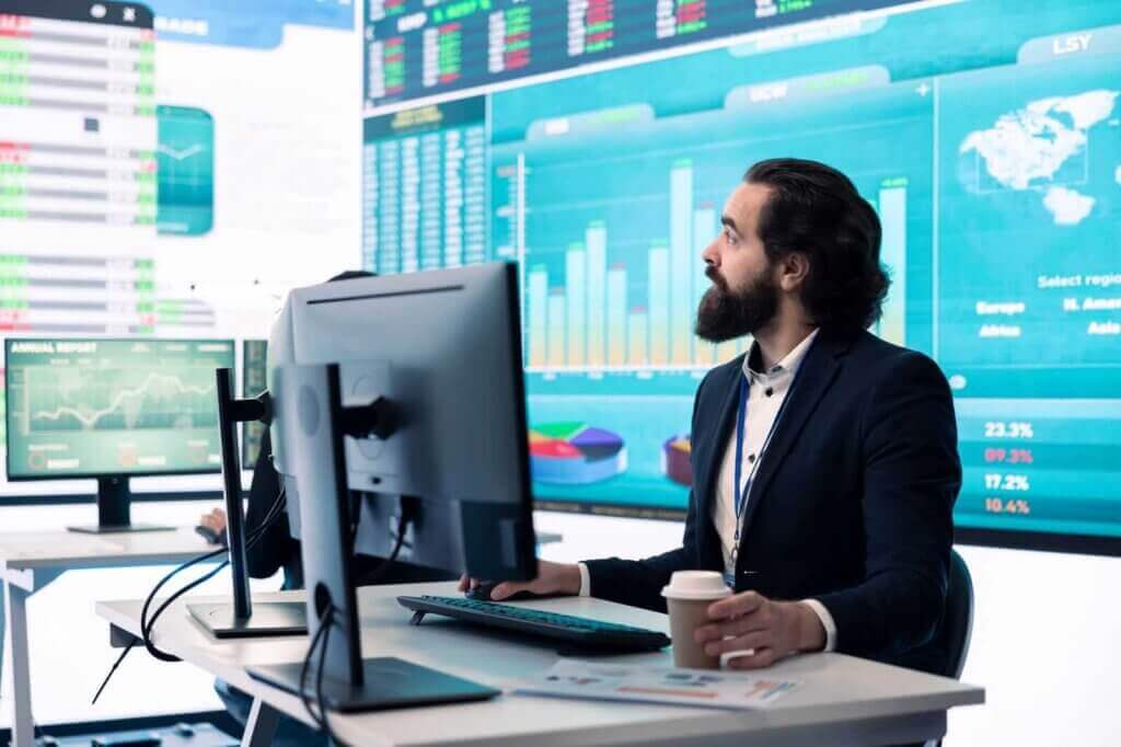 Man in a suit sits at a desk with multiple monitors, analyzing financial data and graphs. He holds a coffee cup in one hand. Large digital screens display charts and statistics behind him.