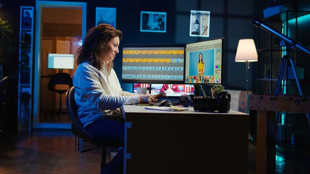 A person is sitting at a desk editing a photo on a computer screen in a dimly lit room with a camera and equipment nearby.