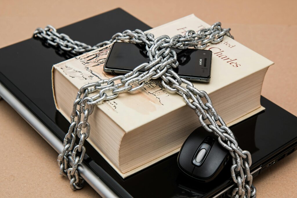 A book, smartphone, and laptop are secured together with chains on a wooden surface, with a computer mouse nearby.