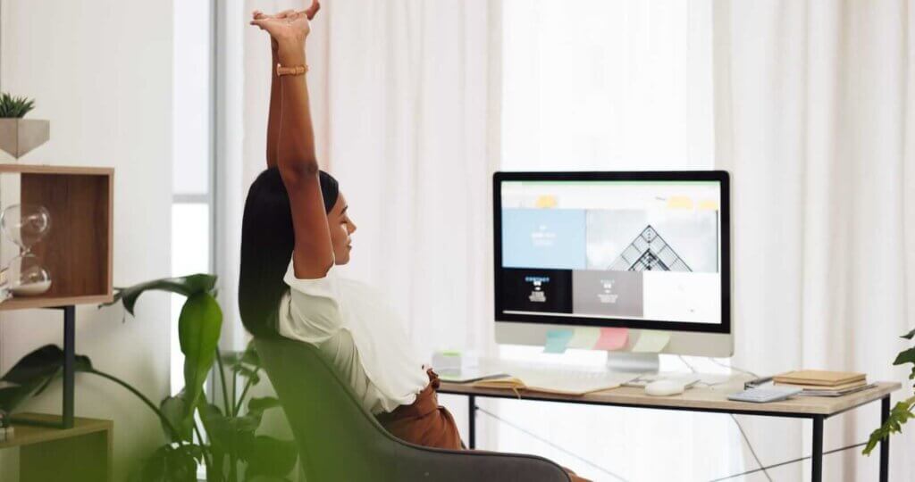 A person stretches with arms raised at a desk in a home office, computer screen visible, surrounded by plants.