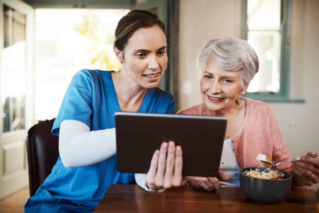 A woman in blue scrubs shows a tablet to an elderly woman eating. They both appear engaged in the content on the screen.