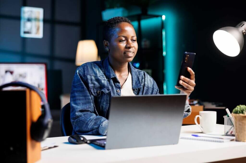 Person in a denim jacket sitting at a desk with a laptop, looking at their smartphone. A lamp, headphones, and a cup are on the desk.