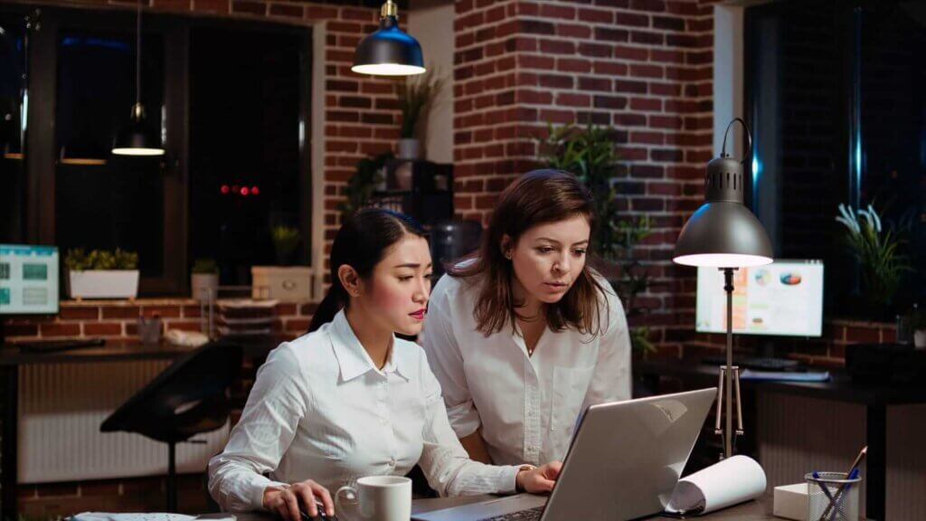 Two women in a brick-walled office, wearing white shirts, work at a desk with a laptop and a cup. They are focused on the screen, with plants and office decor in the background.