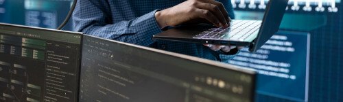 Person in a checkered shirt working on a laptop, with multiple computer monitors displaying code and data in a tech environment.